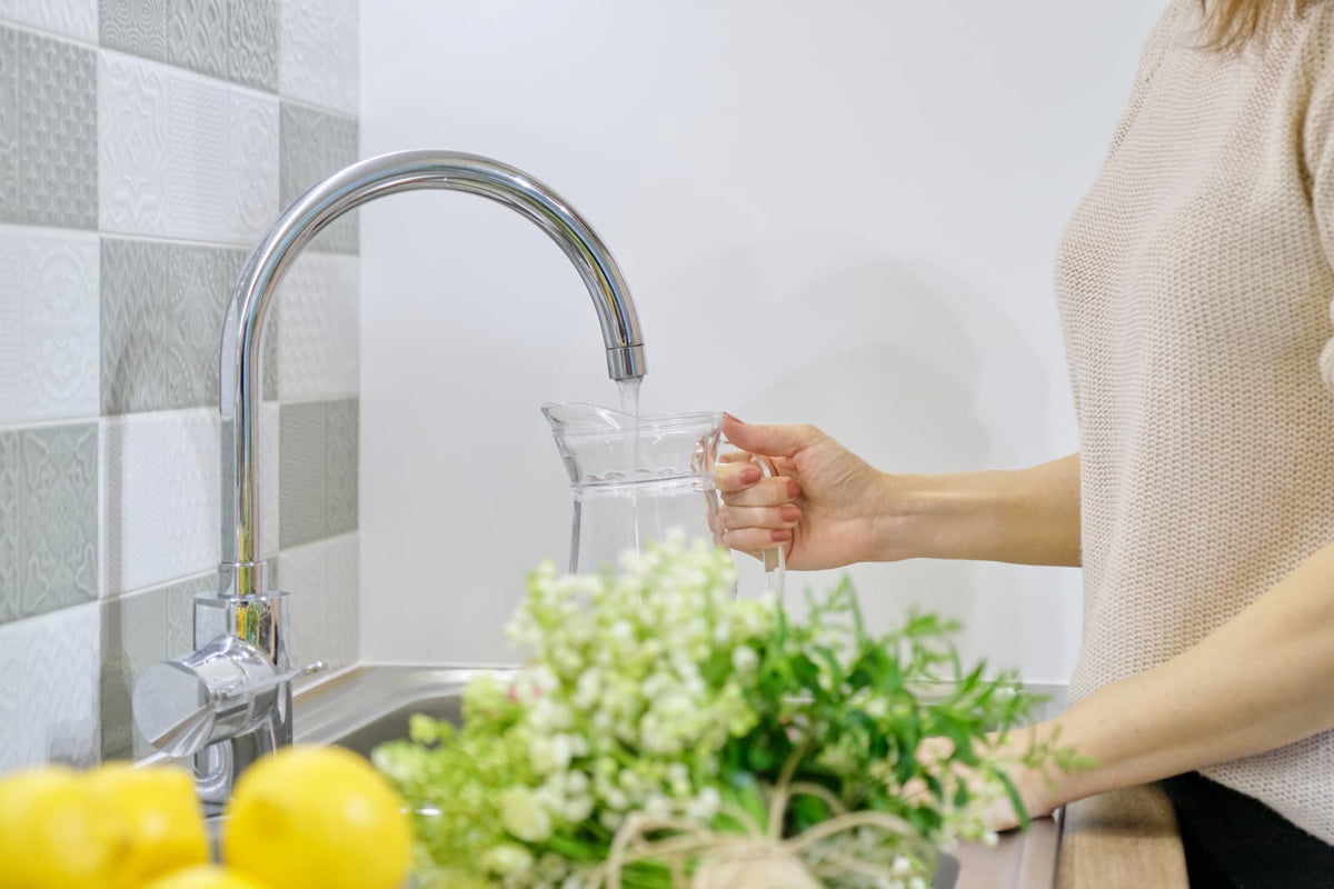 Eine Frau füllt frisches Leitungswasser aus der Küchenarmatur in eine Glaskanne, Trinkwasser mit natürlichem Magnesiumgehalt.