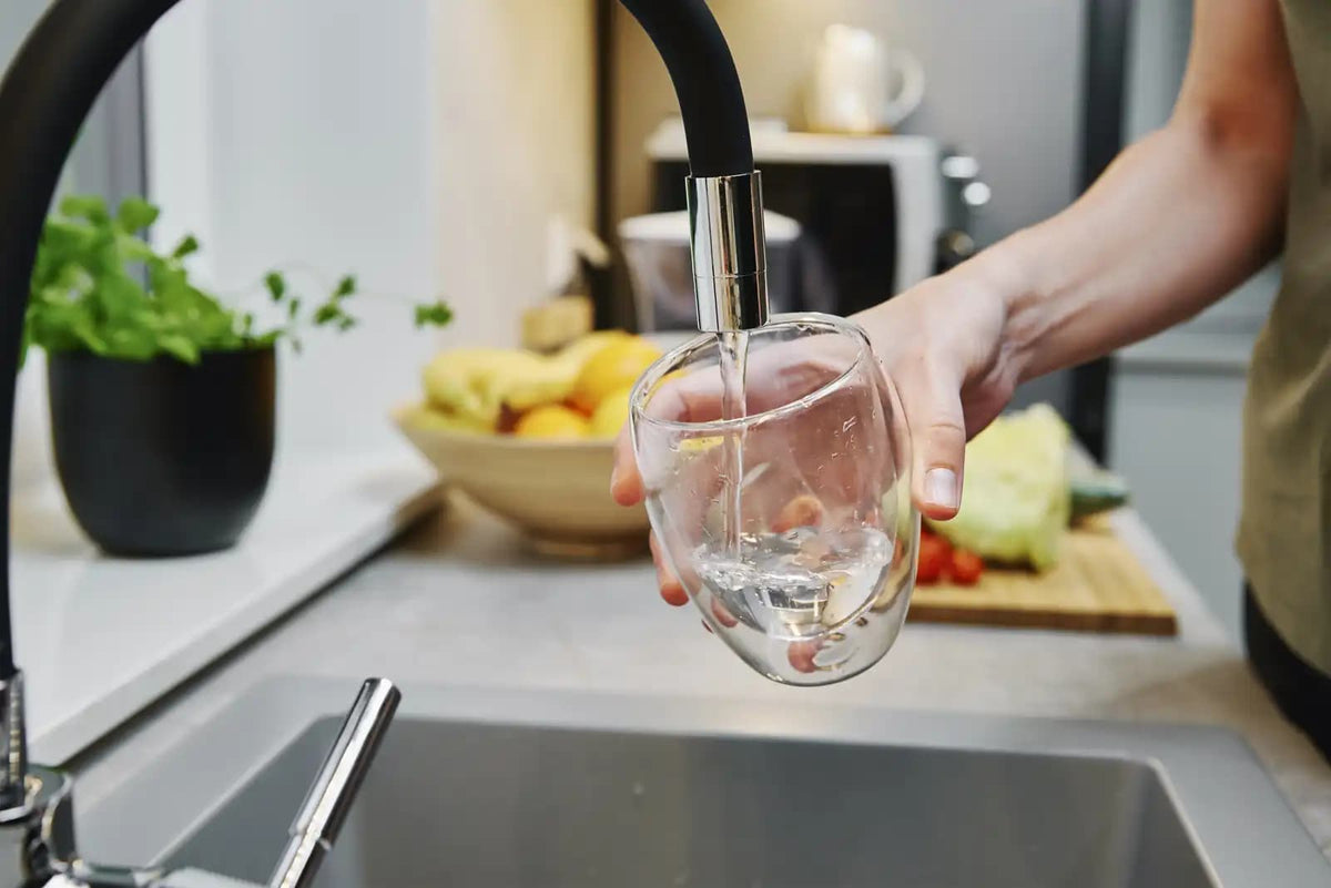 Eine Person füllt ein Glas mit frischem Wasser aus einem modernen Küchenhahn auf einer hellen Küchenarbeitsplatte mit Obst und Grünpflanzen.