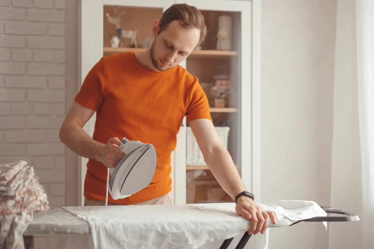 Ein Mann bügelt Kleidung mit einem Dampfbügeleisen zu Hause, ein typisches Haushaltsgerät, für das oft destilliertes Wasser verwendet wird