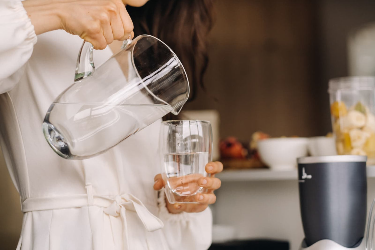 Eine Frau gießt klares, mineralhaltiges Trinkwasser aus einer großen Glaskanne in ein Glas in der modernen Küche.