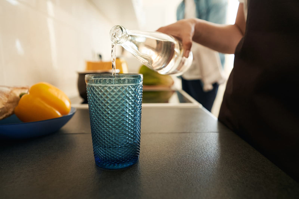 Eine Person gießt klares gefiltertes Wasser aus einer Glasflasche in ein blaues Trinkglas auf der modernen Küchenarbeitsfläche.