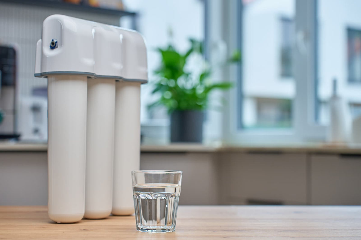 Dreistufiger weißer Wasserfilter auf einem Küchentisch neben einem Glas mit klarem Wasser, im Hintergrund eine moderne Küche mit Fenster und Zimmerpflanze.